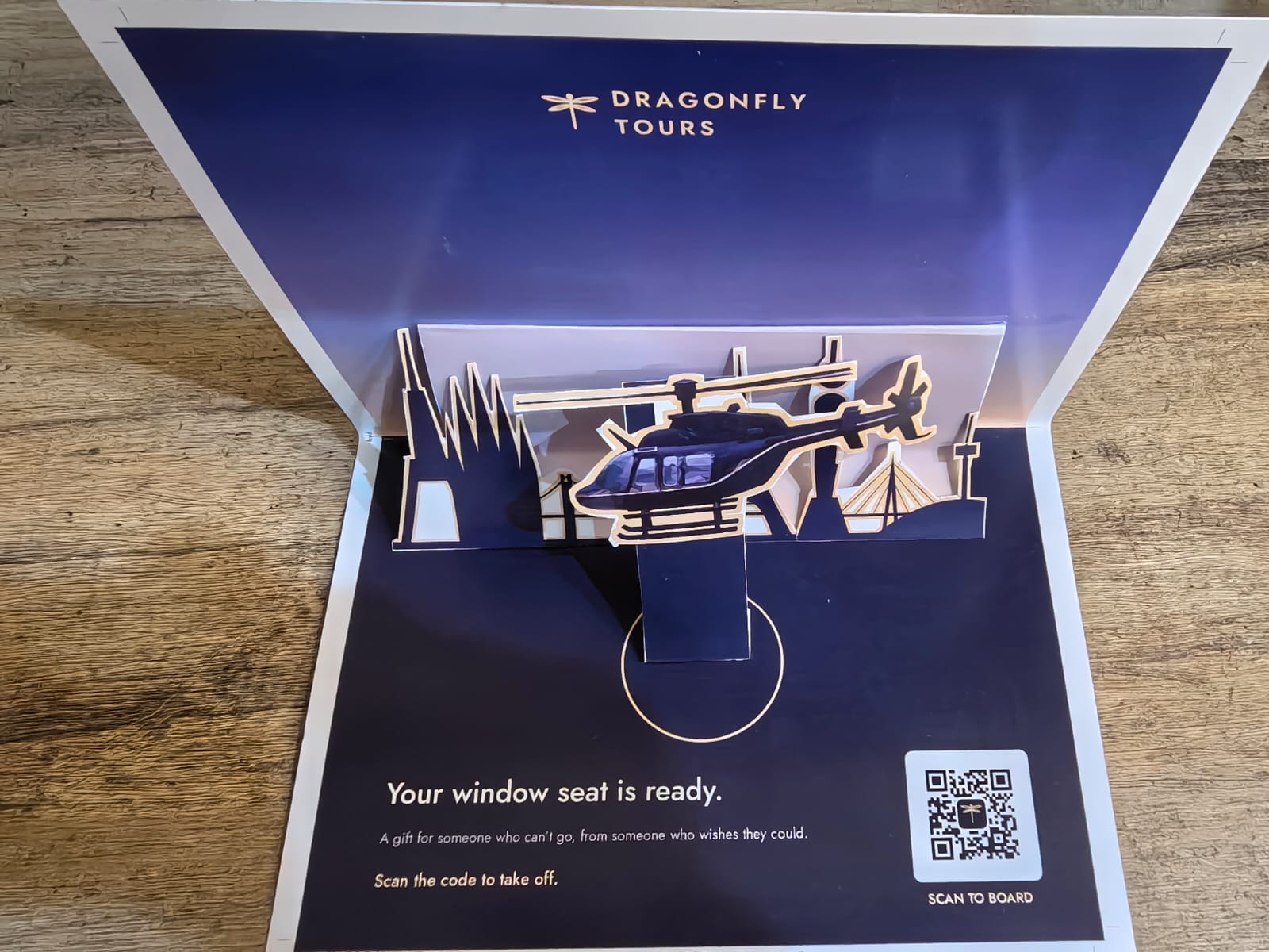 Photograph of the assembled Dragonfly Tours pop-up card with a raised helicopter, layered skyline and scan-to-board QR code